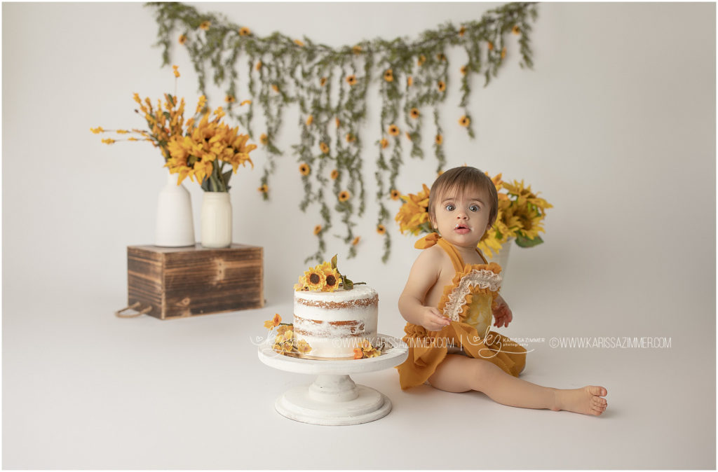 Baby girl dressed in yellow looks surprised to smash her first birthday cake at camp hill photography studio