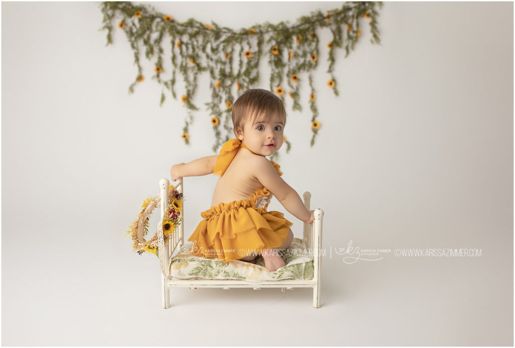 one year old baby girl poses on little bed with sunflowers at her first birthday photoshoot
