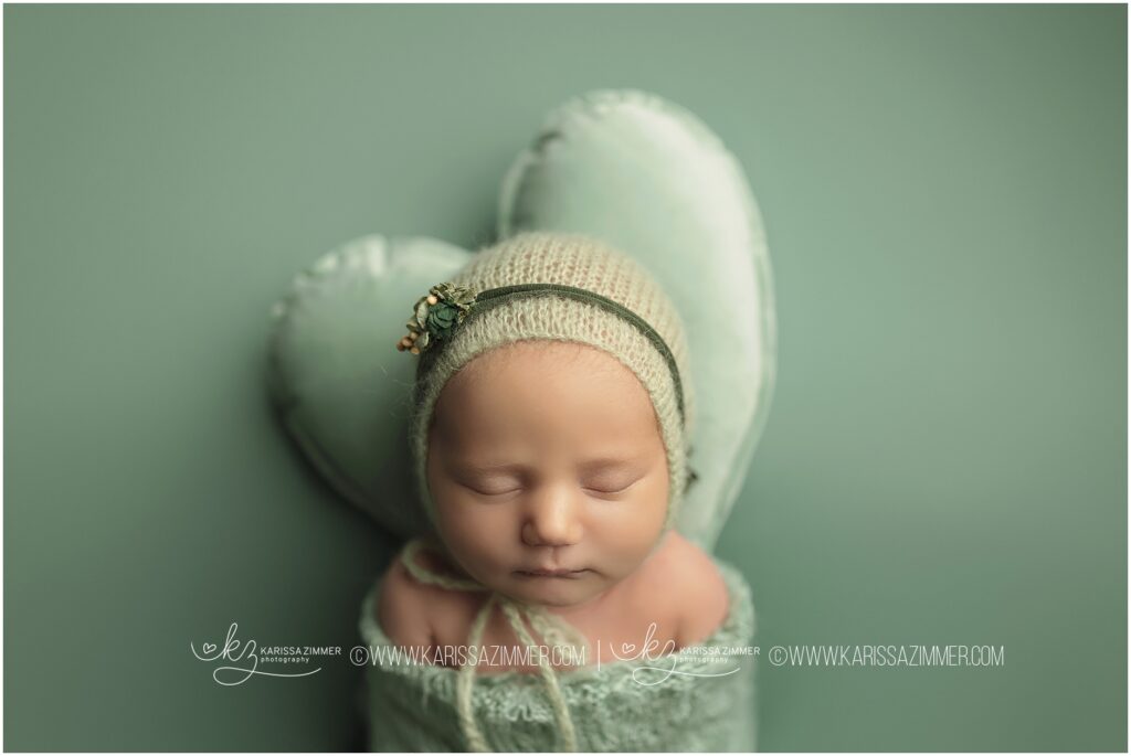 Studio newborn photography of baby girl wearing green and resting head on heart pillow at Karissa Zimmer Photography