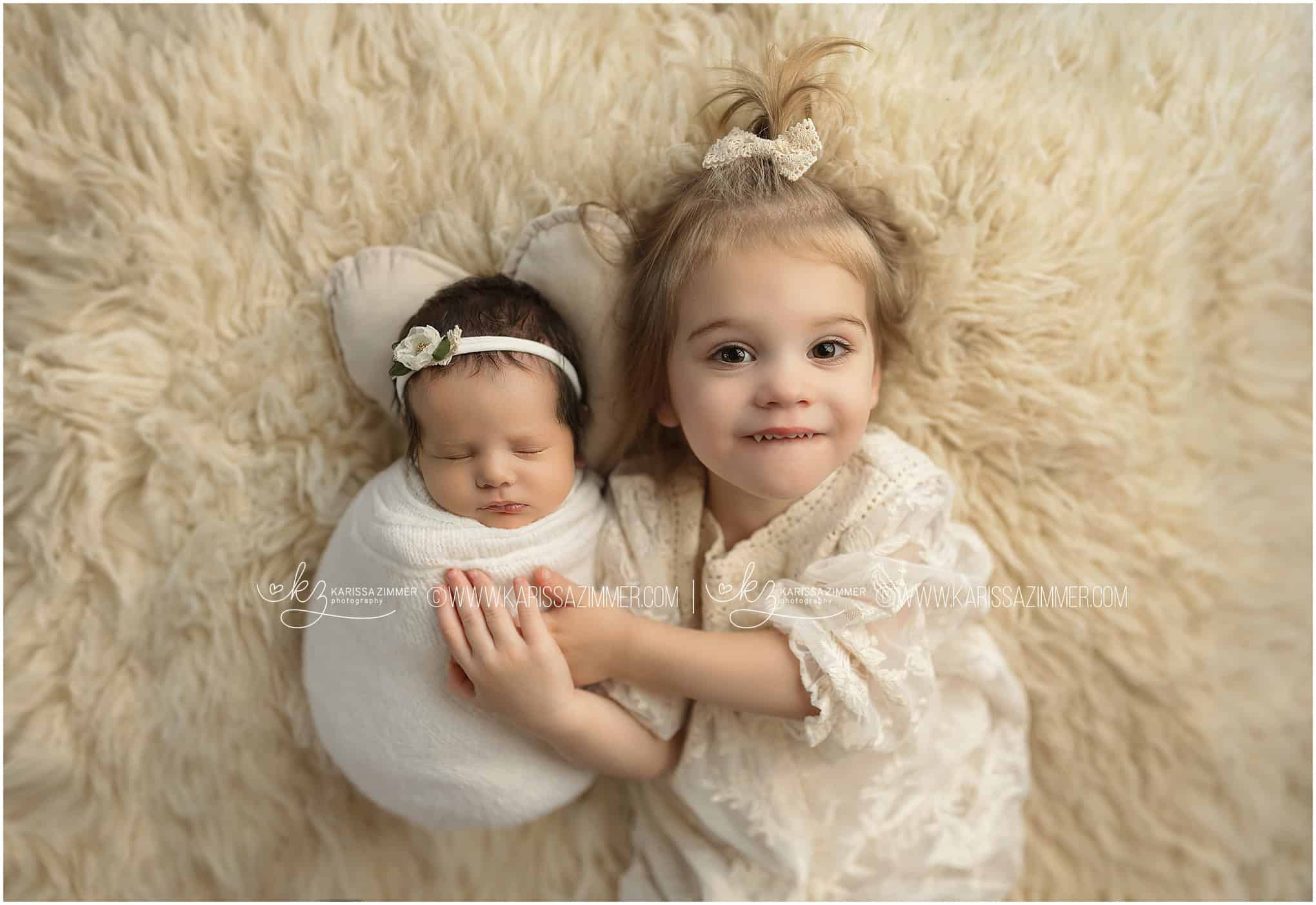 A new big sister poses with her brand new baby sister on all white